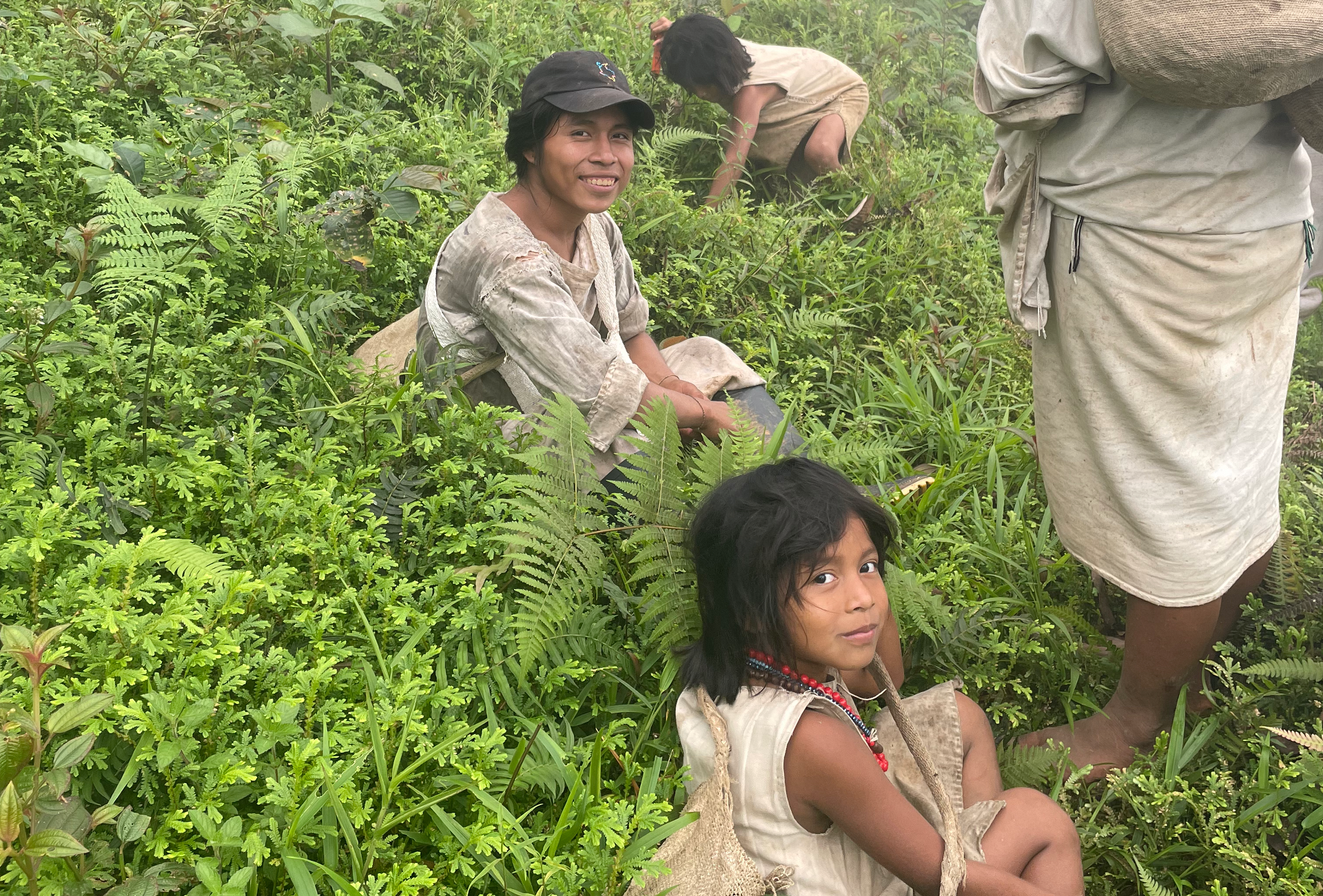 Members of an indigenous community amid dense vegetation, with a child in the foreground wearing traditional beaded necklaces