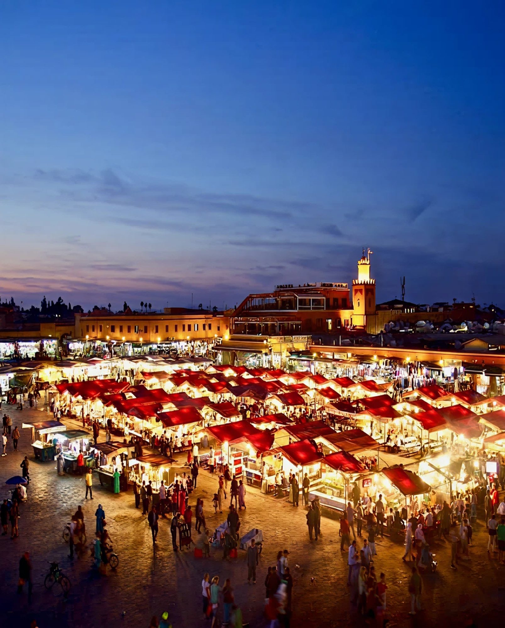 Nighttime view of Jemaa el-Fnaa square in Marrakesh, with illuminated red tents, crowds, and a mosque tower in the background