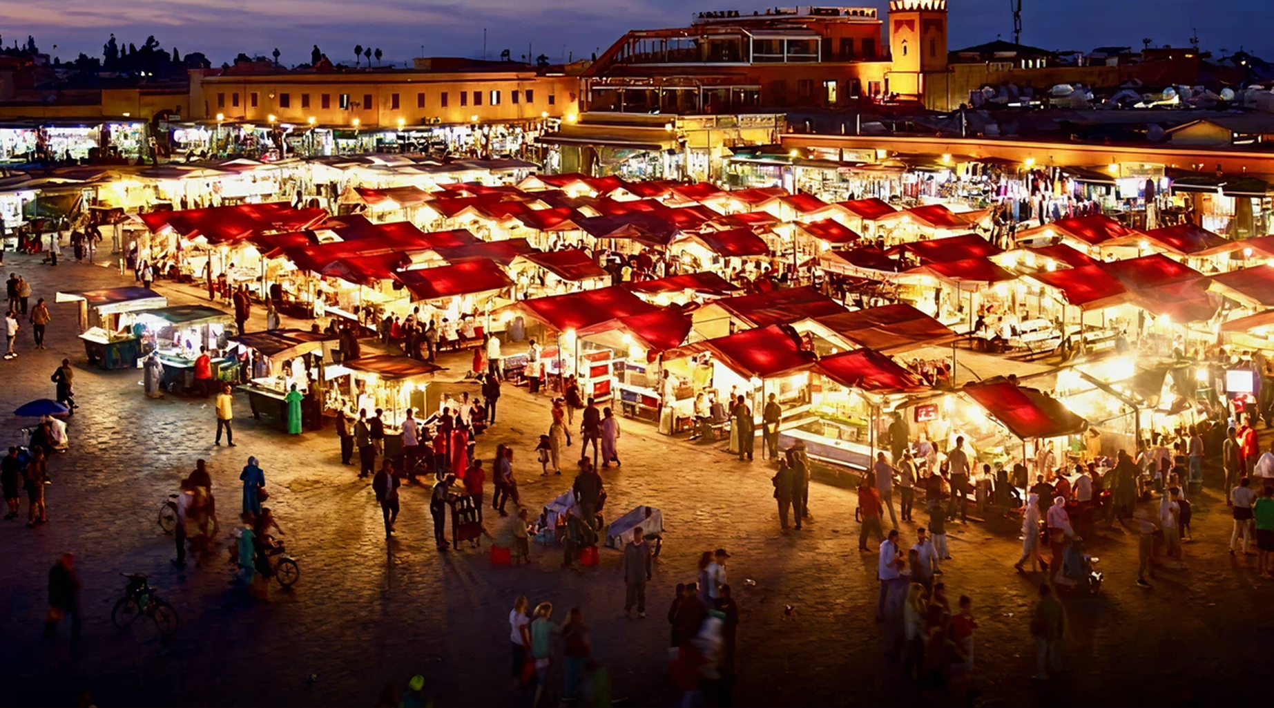 Nighttime view of Jemaa el-Fnaa square in Marrakesh, with illuminated red tents, crowds, and a mosque tower in the background