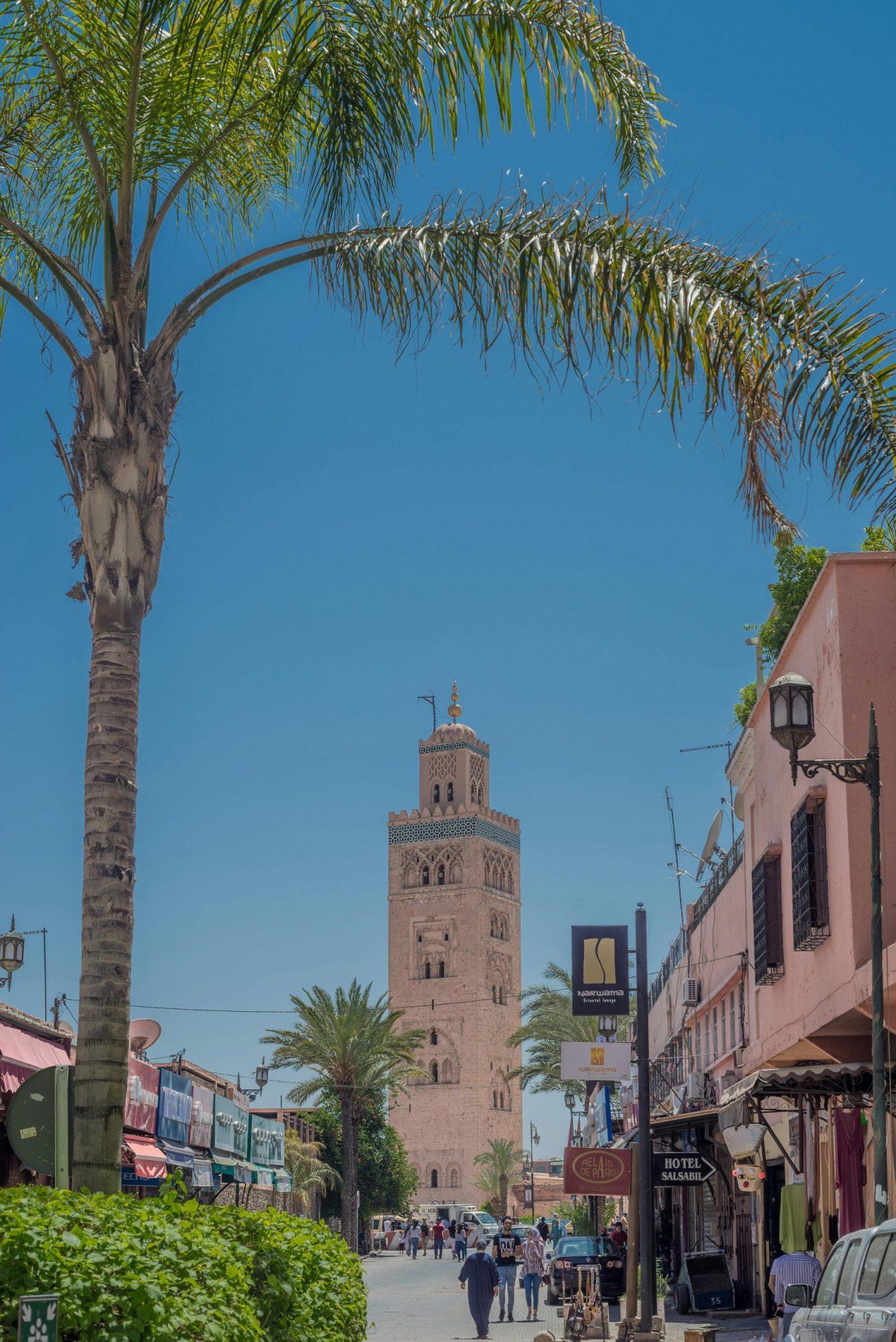 The Koutoubia Mosque tower in Marrakesh, seen from a street lined with palm trees, shops, and hotels